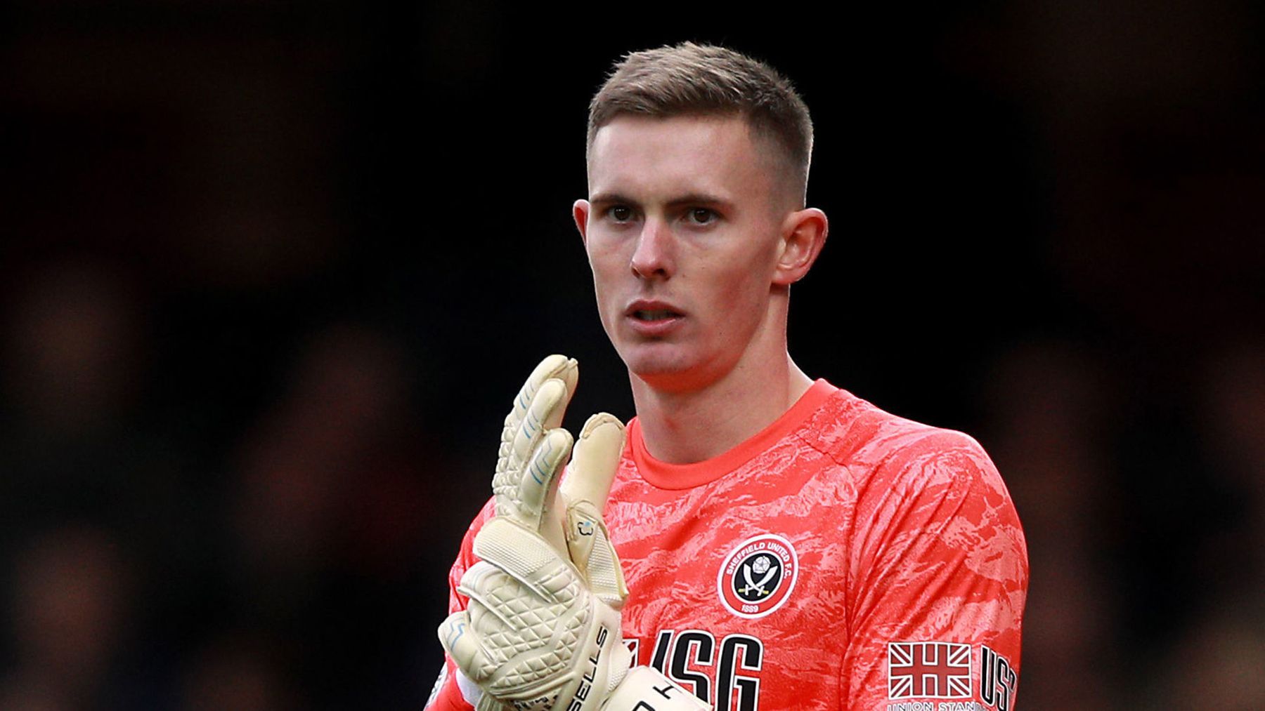  A young male goalkeeper wearing a red and white jersey with gloves is standing in front of a dark background.